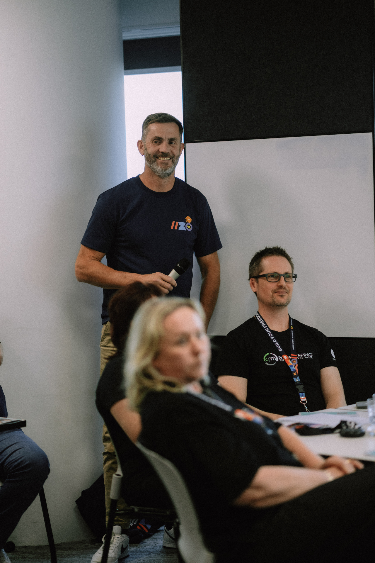 Wize team member smiling with a microphone alongside seated attendees during Day 1